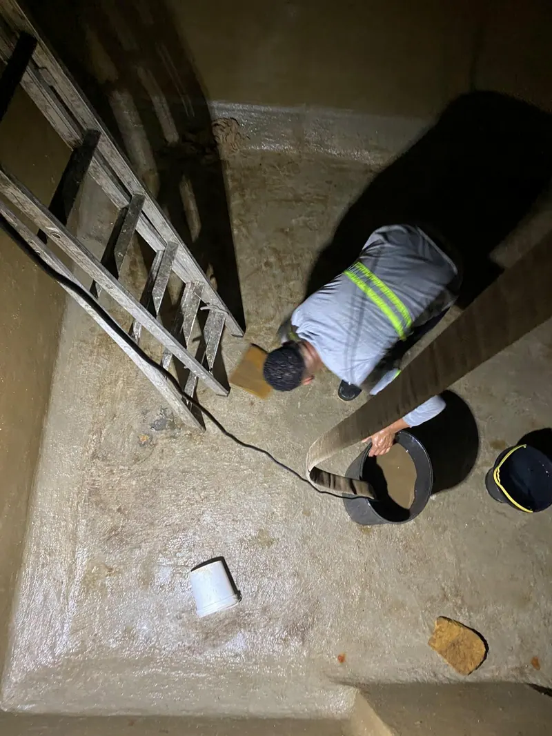 Funcionário de uniforme no fundo da cisterna já limpa, com a escada, a mangueira e os baldes, visto de cima